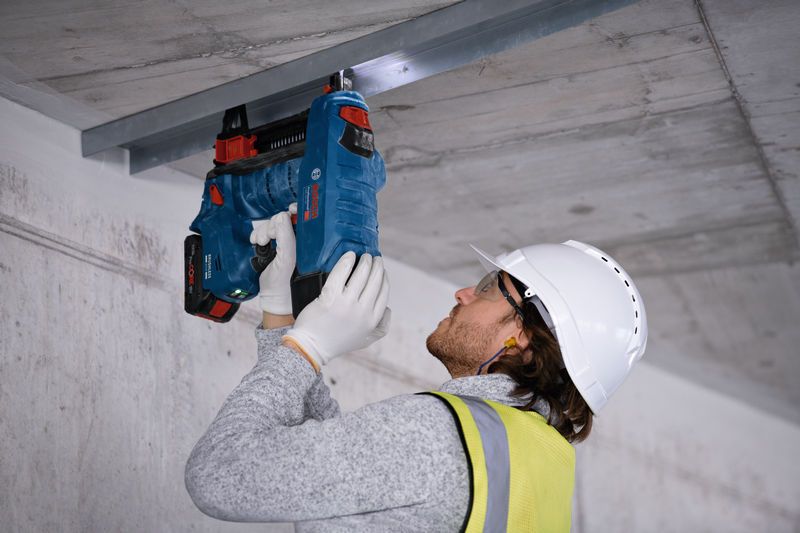 A person wearing safety equipment uses a power tool to fasten a metal frame to a ceiling.