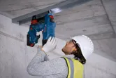 A person wearing safety equipment uses a power tool to fasten a metal frame to a ceiling.