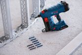 Cordless nail gun placed on a floor beside fastener strips and metal framing studs.