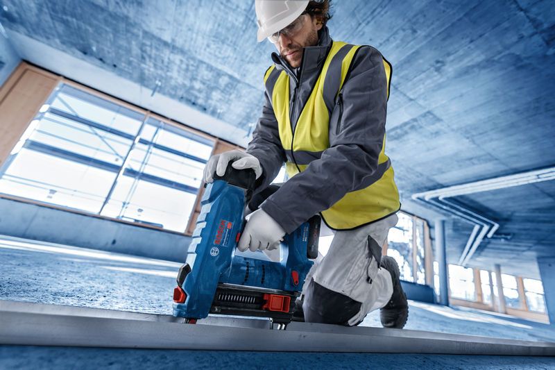 A person wearing safety equipment uses a cordless concrete nailer on a construction floor.