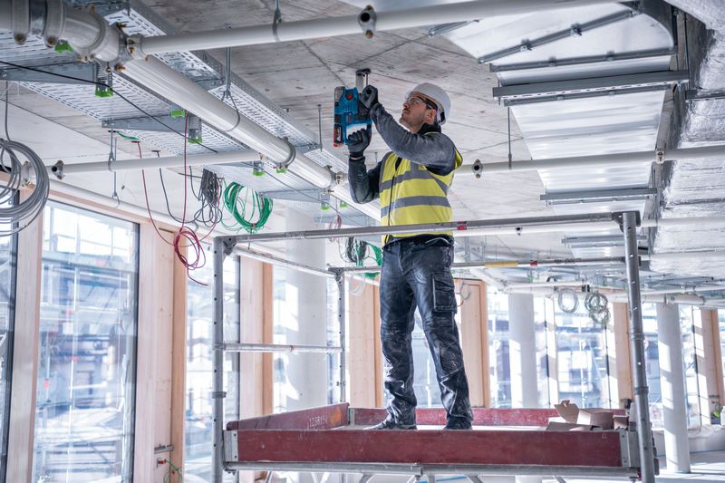 A person wearing safety equipment drills into a concrete ceiling on a construction site.