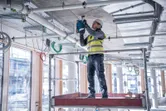 A person wearing safety equipment drills into a concrete ceiling on a construction site.