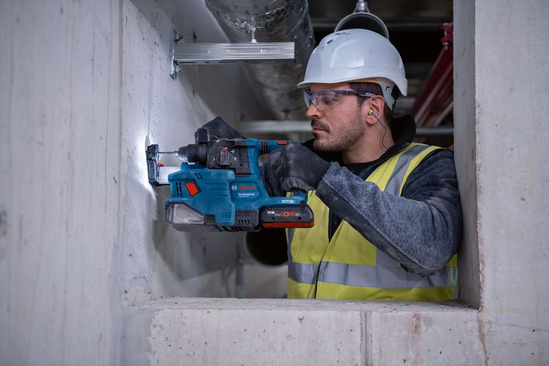 A person wearing safety equipment drills into a concrete wall using a cordless rotary hammer.