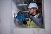 A person wearing safety equipment drills into a concrete wall using a cordless rotary hammer.