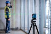 A worker wearing safety equipment aligns a wall frame using a laser leveling tool.
