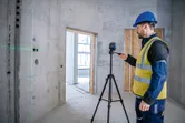 A person wearing safety equipment operates a laser leveling tool on a tripod in a concrete room.