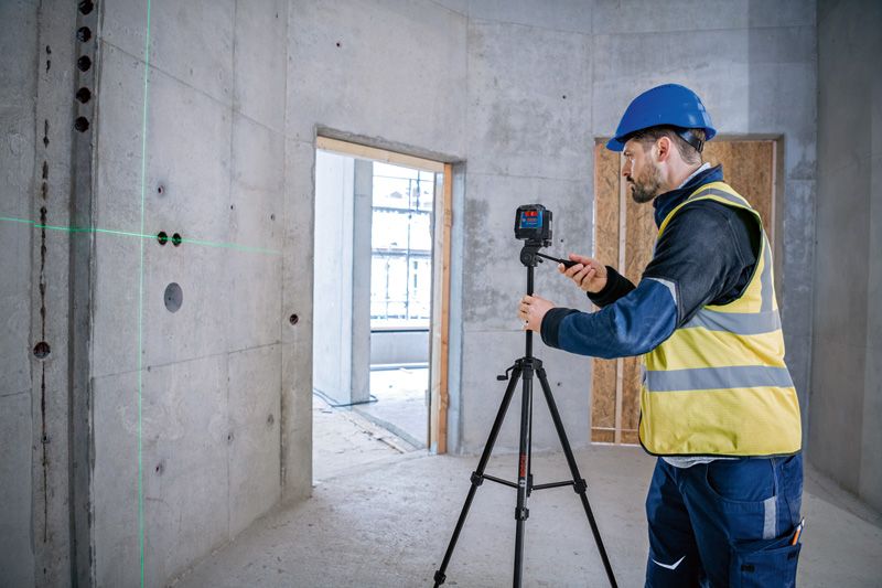 A person wearing safety equipment adjusts a laser leveling tool on a tripod in a concrete room.