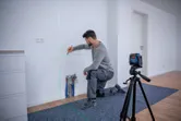 Person kneeling and marking a wall, guided by a laser leveling tool on a tripod.