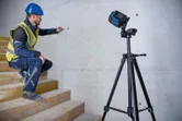 A person wearing safety equipment aligns a stairwell wall using a laser leveling tool on a tripod.