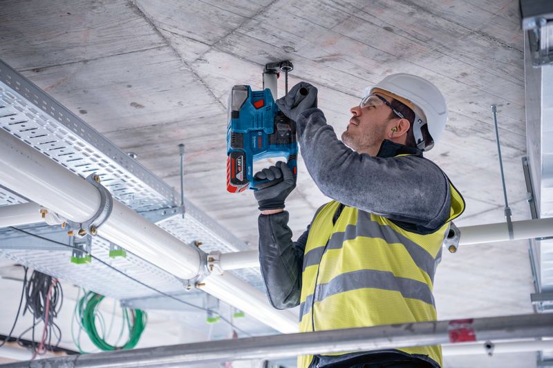 A person wearing safety equipment drills into a concrete ceiling on a construction site.