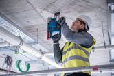 A person wearing safety equipment drills into a concrete ceiling on a construction site.