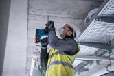 A person wearing safety equipment drills into a concrete ceiling at a construction site.