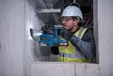 A person wearing safety equipment drills into a concrete wall with a rotary hammer.