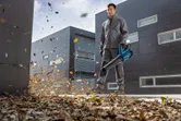 A person wearing safety equipment clears leaves with a cordless leaf blower outside a building.