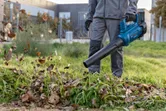 A person wearing safety equipment uses a cordless leaf blower to clear leaves on grass.