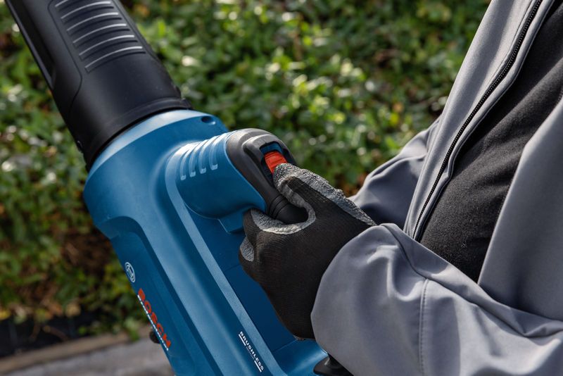 A person wearing safety equipment operates a cordless leaf blower outdoors.