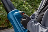 A person wearing safety equipment operates a cordless leaf blower outdoors.