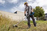 A person wearing safety equipment trims grass with a cordless brushcutter outdoors.
