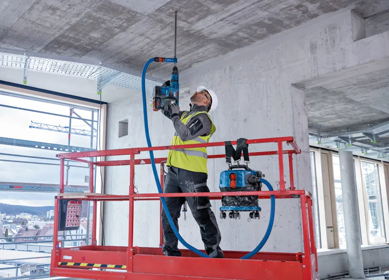 A person wearing safety equipment drills into a concrete ceiling using a cordless dust extractor.
