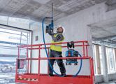 A person wearing safety equipment drills into a concrete ceiling using a cordless dust extractor.