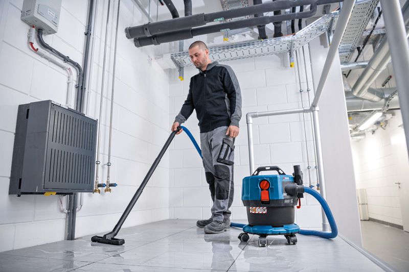 A person wearing safety equipment uses a cordless dust extractor to clean a tiled floor.