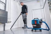 A person wearing safety equipment uses a cordless dust extractor to clean a tiled floor.