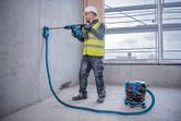 A person wearing safety equipment drills into concrete, cordless dust extractor attached.