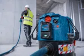 A person wearing safety equipment operates a power tool connected to a dust extractor.