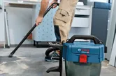 A person wearing safety equipment vacuums dust from a tiled floor in a workspace.