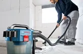A person vacuums water from a concrete floor in a bright unfinished room.
