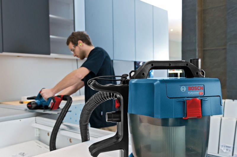 GAS 18V-10 L A person wearing safety equipment uses a power planer on a wooden board near a vacuum.