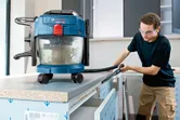 A person wearing safety equipment uses a wet and dry vacuum to clean a workbench.