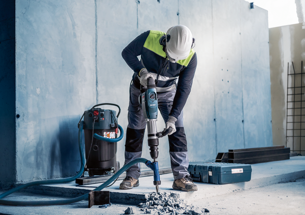 Worker wearing safety equipment uses a demolition hammer to break concrete on a construction site.