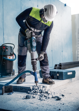 A worker wearing safety equipment drills into concrete with a demolition hammer.
