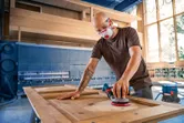 A person wearing safety equipment sands a wooden door with a power sander.