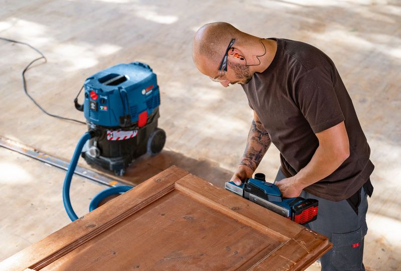 Person wearing safety equipment uses a power planer on a wooden door with a vacuum nearby.
