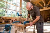 A person wearing safety equipment sands wood on a workbench using a power tool.