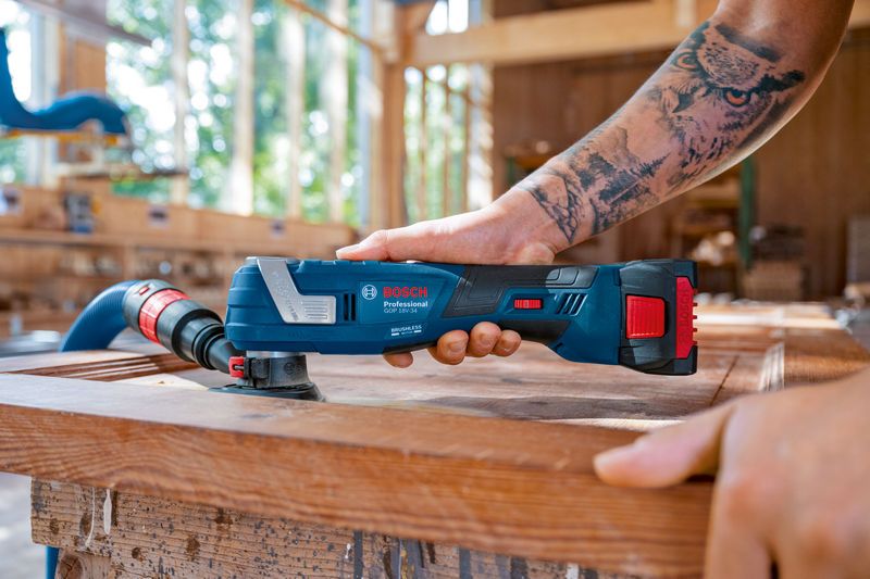 A person sands wood with a cordless multi-tool connected to a dust extraction hose.