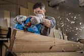 A person wearing safety equipment smooths a large wooden beam with an electric planer.