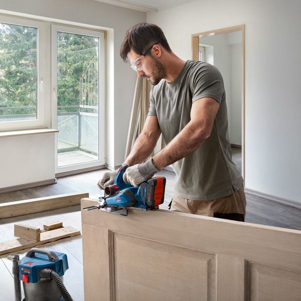 Person wearing safety equipment cuts a wooden door with a power saw indoors.