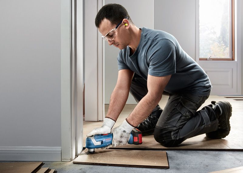 A person wearing safety equipment trims wooden flooring with a cordless multi-tool.
