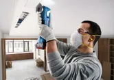 A person wearing safety equipment uses a cordless cutting tool on a ceiling panel.