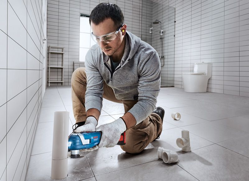 A person wearing safety equipment cuts a PVC pipe with a cordless power tool in a tiled bathroom.