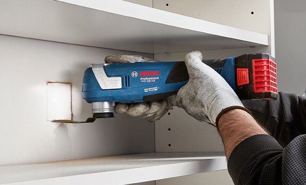 A person wearing safety equipment uses a multi-tool to cut a hole in a shelf.