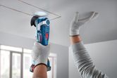 A person wearing safety equipment cuts a ceiling panel with a cordless multi-tool.