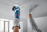 A person wearing safety equipment cuts a ceiling panel with a cordless multi-tool.