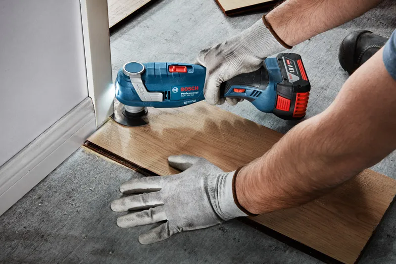 A person wearing safety equipment cuts flooring near a door using a cordless multi-tool.