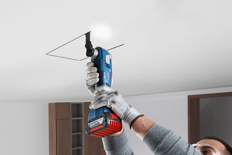 A person wearing safety equipment cuts a square opening in the ceiling with a cordless power tool.