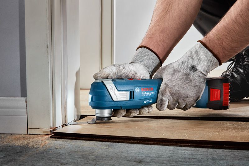 A person wearing safety equipment trims flooring near a door frame with a multitool.