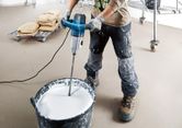A person wearing safety equipment mixes paint with a powered stirring mechanism.